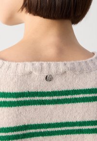 Cheveux courts bruns au niveau du cou et des épaules, portant un pull en tricot blanc avec des rayures vertes et une petite étiquette métallique ronde « MOS MOSH » au col.