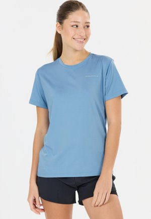 Junge Frau mit Pferdeschwanz, die ein hellblaues Sportshirt mit kurzen Ärmeln und schwarze Sportshorts trägt, lächelt und zur Seite schaut.