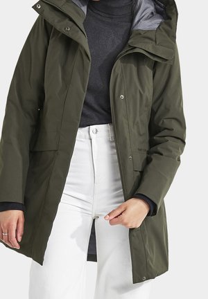 Personne portant une veste d'hiver à capuche vert olive sur un col roulé gris foncé et un pantalon blanc, ajustant le devant de la veste avec la main droite.