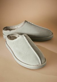 Des chaussons en suede gris clair avec un intérieur doux et moelleux. Dotés d'un bout arrondi et de coutures décoratives le long des coutures. Semelle plate.