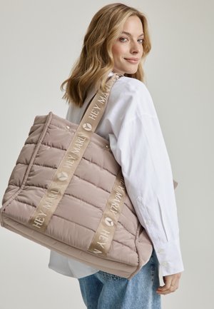 Light beige quilted tote bag with padded texture; features wide, branded tan straps and subtle hardware accents. Worn over the shoulder.
