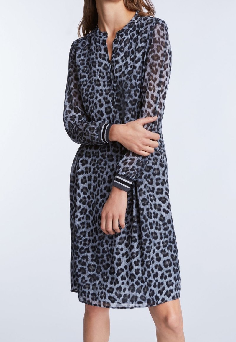 Kobieta w szarej sukience o długości do kolan, z długim rękawem, pokrytej czarnymi plamami w panterkę oraz w paskowane mankiety, stojąca z założonymi rękami.