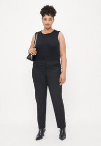 Black sleeveless top, fitted black trousers, and black ankle boots. Model holds a small black bag. Simple design, solid colors.