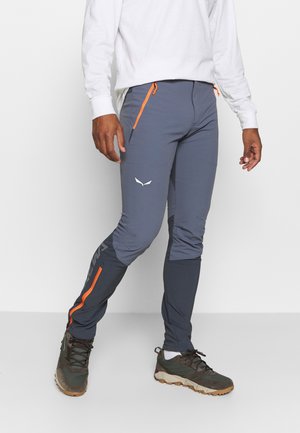Mann trägt graue Sporthose mit orangen Reißverschlüssen und dunkle Wanderschuhe, steht vor einem einfarbigen weißen Hintergrund.