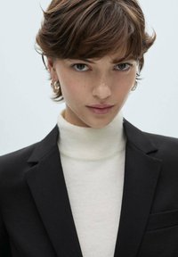 Black blazer over a cream turtleneck sweater. Short brown hair, silver hoop earrings. Close-up of face, neutral expression, light background.