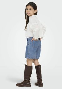 Witte lange mouwen top met kantaccenten, gecombineerd met denim shorts en bruin leren laarzen tot de knie. Kind staat met handen in de zakken.