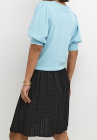 Kvindelig iført en lys blå kort pufærmet sweater og en sort plisseret nederdel, med et guldbelagt armbånd på håndleddet, foran en ensfarvet baggrund.