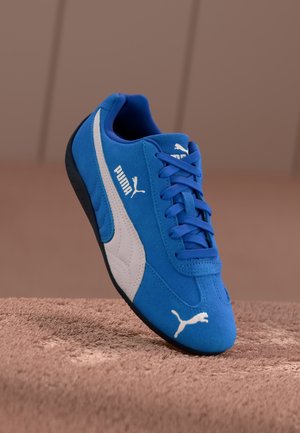 Baskets Puma bleues avec logo blanc et bande, lacets bleus et semelle noire reposant sur une surface texturée marron.