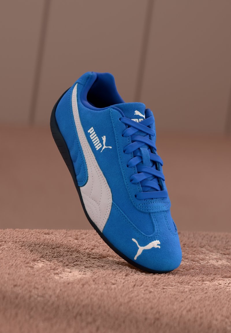 Baskets Puma bleues avec logo blanc et bande, lacets bleus et semelle noire reposant sur une surface texturée marron.