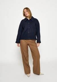 Tumesinist ülikõrge jakk terava kraega ja nelja suure nupuga, kombineeritud laiade pruuni värvi pükstega ja koorevärvi kontsakingadega.