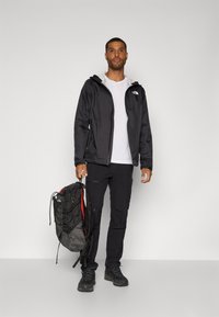 Schwarze wasserdichte Jacke mit Kapuze, weißes T-Shirt, schwarze Hose und robuste schwarze Stiefel. Hält einen schwarz-roten Rucksack mit verstellbaren Trägern.