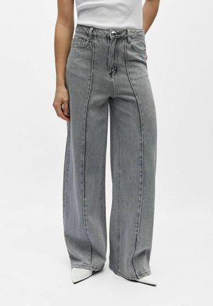 Ženska nosi sive kavbojke z visokim pasom in širokimi hlačnicami z detajli sprednjih šivov ter belimi čevlji z ostro konico, kombinirane z belim brezrokavnikom.