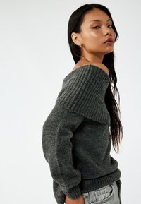 Grå off-the-shoulder sweater med ribbet halsudskæring, lange ærmer og en struktureret overflade. Har en tætsiddende kant og en afslappet silhuet.