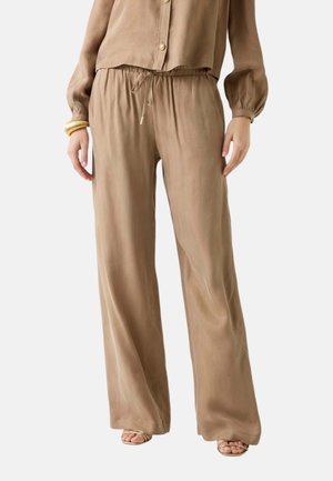 Pantaloni beige dal taglio ampio con vita elasticizzata e coulisse, indossati con una camicia beige abbottonata coordinata e sandali con punta aperta.