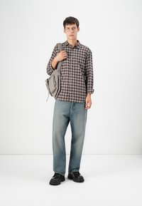 Plaid button-up shirt in brown and white, light blue jeans, black chunky shoes, carrying a grey bag, standing against a plain white background.