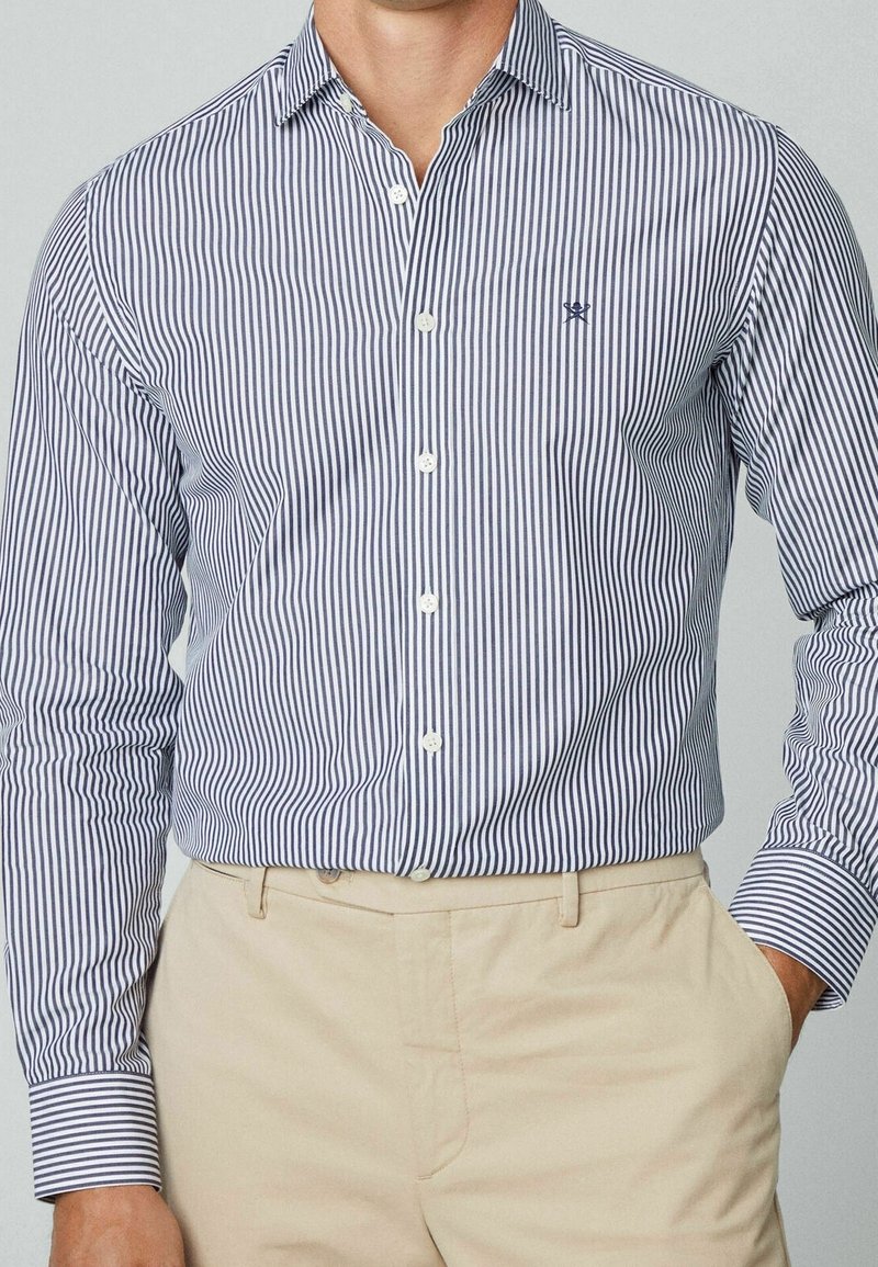 Uomo che indossa una camicia azzurra e bianca a righe verticali, infilata in pantaloni beige, con una mano in tasca.