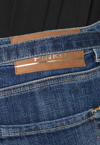Jeans de denim con un lavado azul oscuro, que presentan costuras en contraste y una etiqueta de cuero color marrón claro con "PINKO" grabado en plata en la cintura.