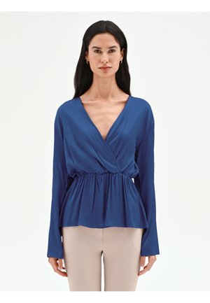 Femme aux longs cheveux foncés portant un chemisier bleu à manches longues, croisé avec une taille froncée, et un pantalon beige, debout devant un fond uni.