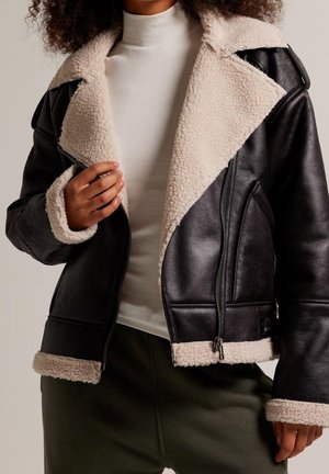 Person iført sort læderjakke med cremefarvet fleecefor, hvid ribbet rullekrave og mørkegrønne bukser, der holder om jakkereverset.