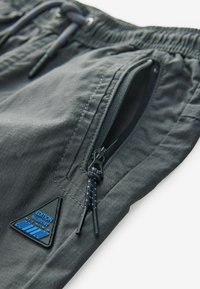 Shorts gris en tissu léger avec une taille élastique, dotés de deux poches zippées et d'un logo en triangle bleu et noir.