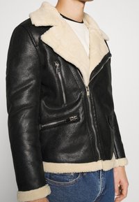 Czarna skórzana kurtka z beżowym futrzanym kołnierzem i wykończeniem. Posiada srebrne kieszenie na zamek błyskawiczny oraz dopasowany krój. Teksturowana powierzchnia.