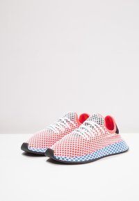 Des chaussures de sport en maille avec un dessus blanc, des accents rouges et une semelle à motif bleu. Les caractéristiques incluent des lacets et un col rembourré.