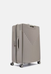 Valigia rigida beige con scanalature verticali, maniglia superiore, chiusura a zip e quattro ruote girevoli per una facile mobilità.