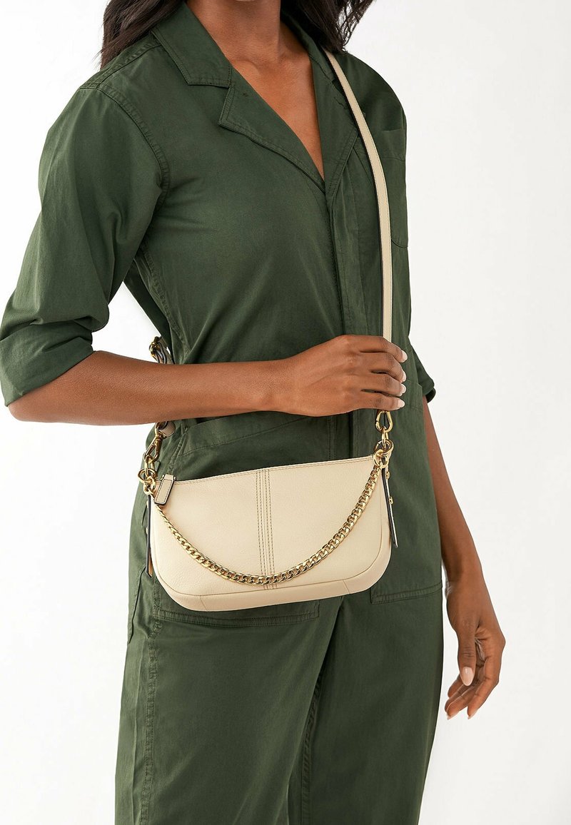 Beżowa skórzana torebka crossbody o zaokrąglonym kształcie, z łańcuchem jako akcentem i regulowanym paskiem, trzymana przez osobę w zielonym stroju.