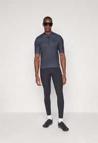 Haut de sport à manches courtes, de couleur bleu marine, avec une fermeture éclair à l'avant, associé à des leggings noirs ajustés. Le mannequin porte des lunettes de soleil noires et des chaussures.