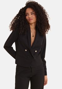 Blazer nero doppiopetto con bottoni dorati, vestibilità sartoriale, rever a punta e texture liscia, abbinato a pantaloni coordinati.