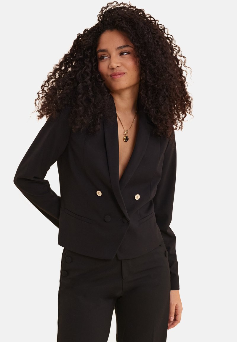 Blazer nero doppiopetto con bottoni dorati, vestibilità sartoriale, rever a punta e texture liscia, abbinato a pantaloni coordinati.