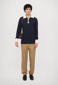 Navy pitkähihaista polo, jossa on kerma väri kauluksessa, beige housut, ruskeat loafers-kengät ja ruskea olkalaukku. Etupuolella on vetoketju ja logopatch.