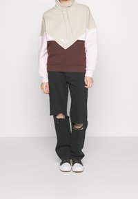 Hoodie i beige, rosa och bruna färgblock; har en logotyp på bröstet. Bärs med slitna svarta jeans och sneakers.