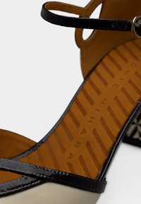 Μαύρο και μπεζ σανδάλι με γυαλιστερό φινίρισμα, που διαθέτει λεπτό λουράκι στον αστράγαλο και ανάγλυφη σόλα, διακοσμημένο με λογότυπο της μάρκας στην εσωτερική σόλα.