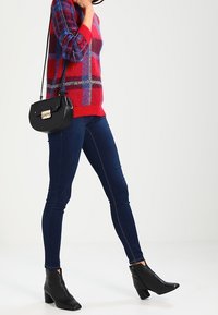 Frau in rotem gemustertem Pullover, dunkelblauen Skinny Jeans, schwarzen Stiefeletten und einer schwarzen Umhängetasche, die vor einem schlichten Hintergrund steht.