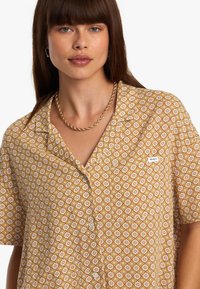 Chemise à manches courtes de couleur marron clair avec un motif floral ; elle dispose d'une fermeture à boutons, d'un col et d'une étiquette logo RVCA sur la poitrine.