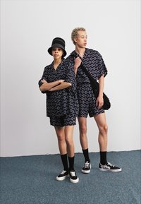 Dos personas llevan camisetas de manga corta y pantalones cortos a juego, con un patrón en azul marino y óvalos de color azul claro. Los accesorios incluyen un sombrero de cubo negro y bolsos.