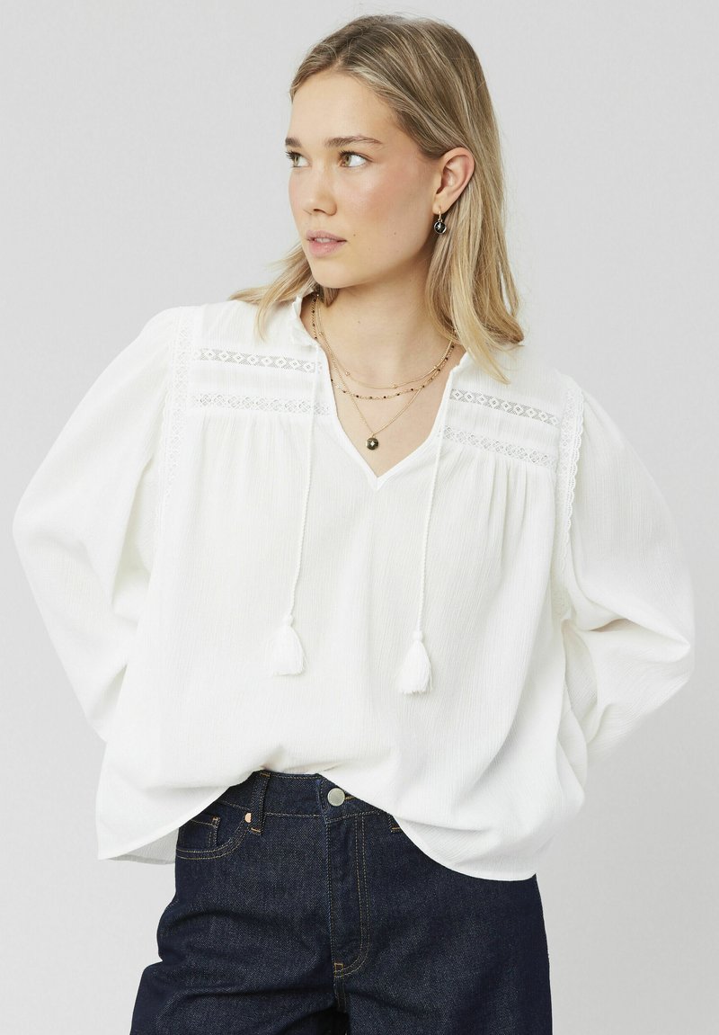 Blouse blanche texturée avec un col en V, ornée d'accents en dentelle et de liens ajustables avec des pompons, assortie à un jean en denim foncé.