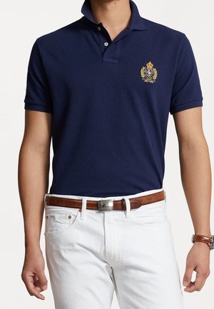 Marineblaues Polohemd aus Baumwolle, mit klassischem Kragen, kurzen Ärmeln und einem goldenen, gestickten Logo auf der linken Brust. Getragen mit weißen Hosen.