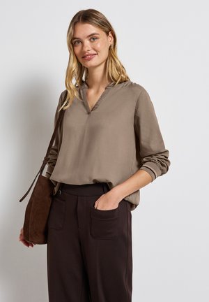 Blouse marron clair avec un col en V et des manches longues. Associée à un pantalon marron foncé avec des poches visibles. Texture de tissu simple et lisse.