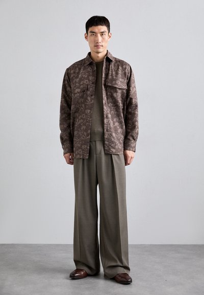 Jachetă cu model maro peste un pulover verde, asortată cu pantaloni bej largi și pantofi maro. Materiale netede și un design ajustat.