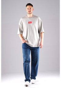 Gray oversized T-shirt with a red logo patch, paired with blue denim jeans, and white sneakers, set against a neutral background.