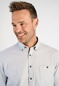 Uomo sorridente con capelli corti e a punta che indossa una camicia grigia chiaro con bottoni e una tasca sul petto, che guarda leggermente a sinistra.