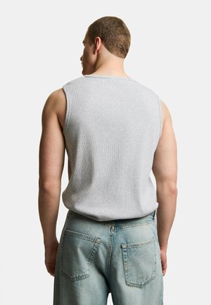 Mężczyzna zwrócony tyłem, ubrany w jasnoszary bezrękawnik i jasnoniebieskie jeansy, na jednolitym białym tle.