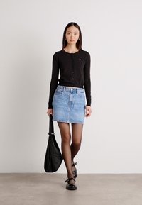Črn dolgrokavnik s gumbi, svetlo modra denim krila, črne pajkice in črne ravne čevlje. Drži črno torbico.