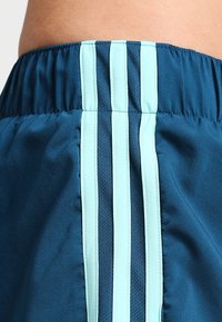 Pantalones cortos deportivos en color verde azulado con una cinturilla elástica y tres rayas verticales de color azul claro a lo largo de la costura lateral. Textura de tela suave.