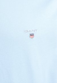 Jasno niebieska koszulka bawełniana z haftowanym logo "GANT" nad wzorem tarczy z gwiazdami i paskami na klatce piersiowej. Gładka tekstura.