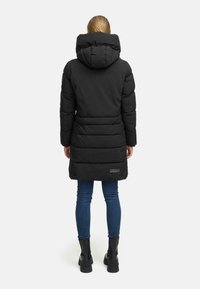 Black padded coat with a hood, featuring vertical stitching, a waistline accent, and a logo patch on the lower back. Worn with jeans and boots.