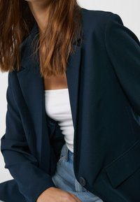 Blazer azul marino con un corte ajustado, hombros estructurados, cierre de un solo botón y solapas con muesca. Textura de tela suave, combinado con una blusa blanca.