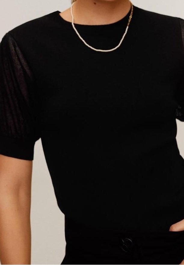 Haut noir avec de courtes manches bouffantes transparentes, encolure arrondie et tissu texturé. Détail d'un délicat collier de perles.